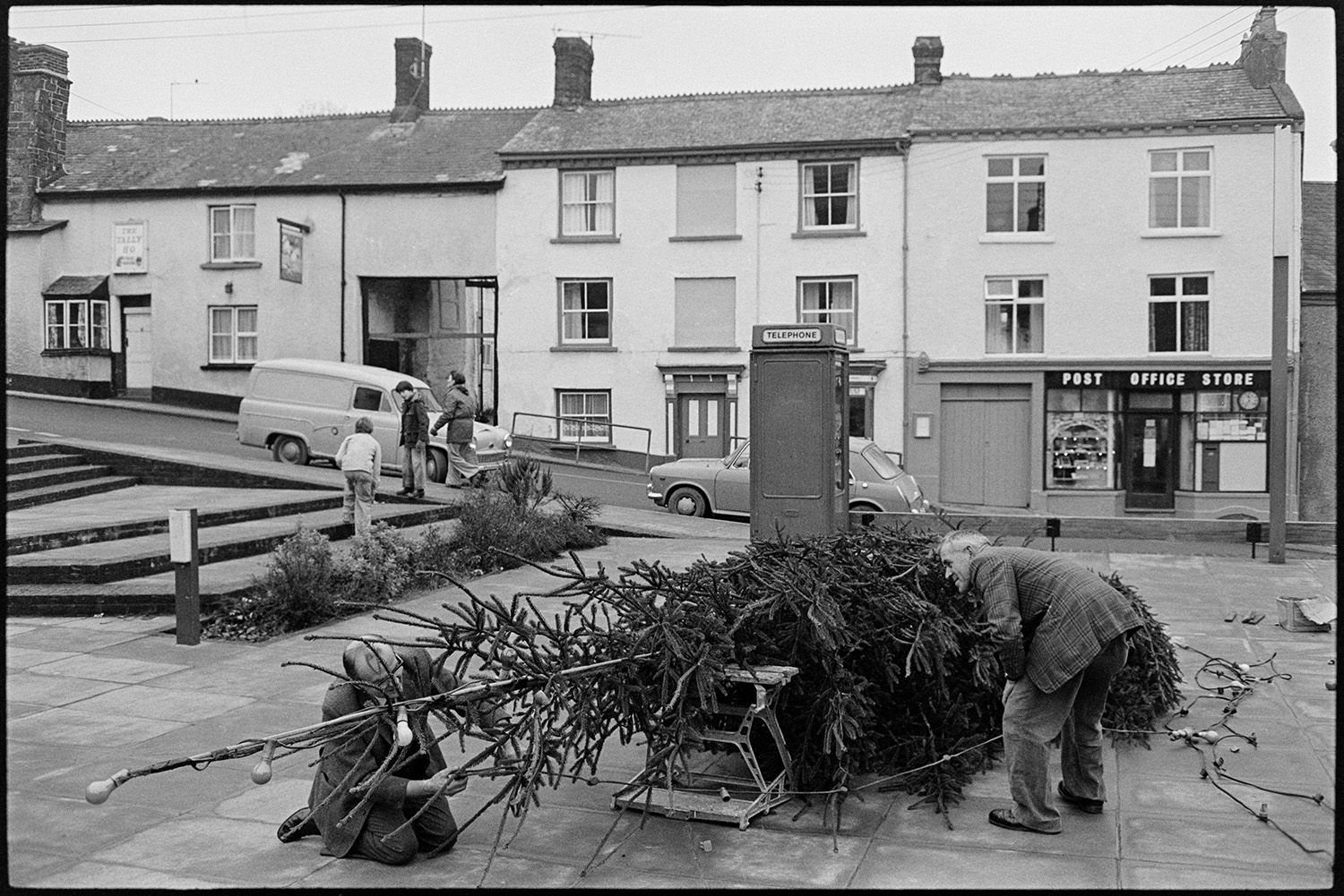 Putting up Christmas tree in town centre. 
[Two men putting up a Christmas tree in the town centre at Hatherleigh, opposite the Post Office and next to a telephone box. In the background people are walking long the street past a parked car and van.]
