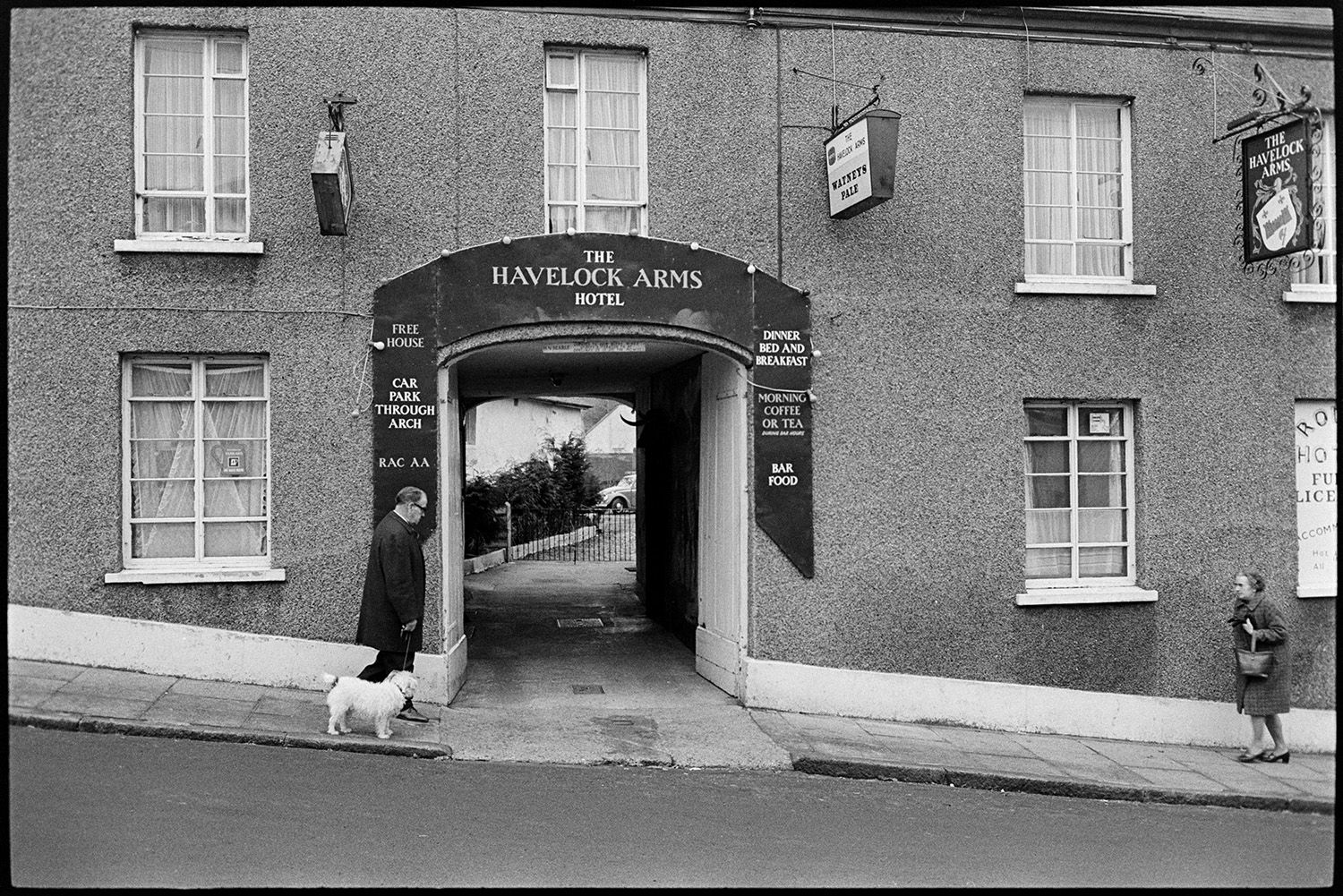 Front of pub, unusual sign. 
[The arched entrance to the Havelock Arms Hotel in Market Street, Hatherleigh. People are walking past the entrance, including a man walking a dog.]