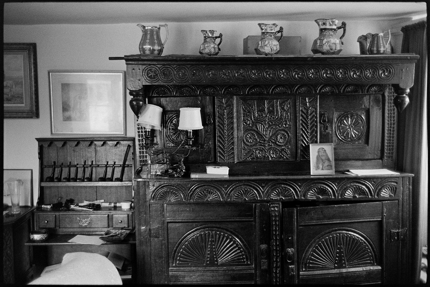 Furniture, carved dresser, jugs. 
[A room in a house in Brightly, Dolton with a carved wooden dresser and a writing desk. A lamp and jugs are displayed on the dresser. Pictures are also hung on the wall.]