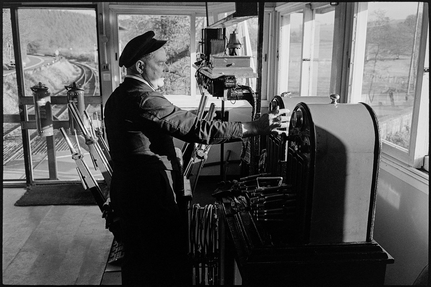 Signal box, signalman operating equipment and handing baton to train driver. 
[Jimmy Hughes, signalman, operating equipment in the signal box at Eggesford Station. The railway tracks can be seen outside the signal box.]