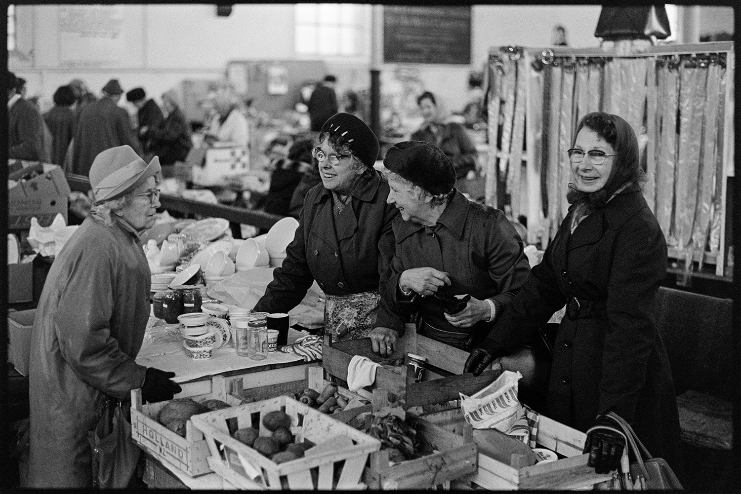 Pannier market stalls and women stallholders, books, vegetables. 
[A market trader and customers talking over a vegetable stall at Bideford Pannier Market.]