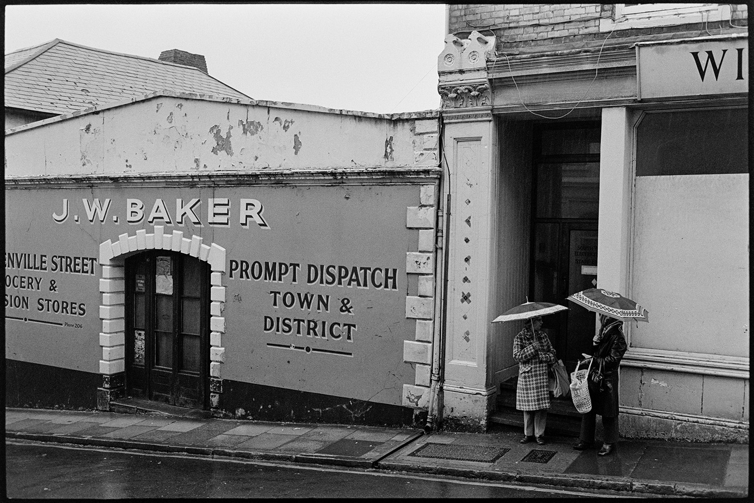 Sign outside Grocery stores in street with passers by. 
[Two people taking in Buttgarden Street, Bideford outside J W Baker, Grocery Stores, in the rain. They are both holding umbrellas.]