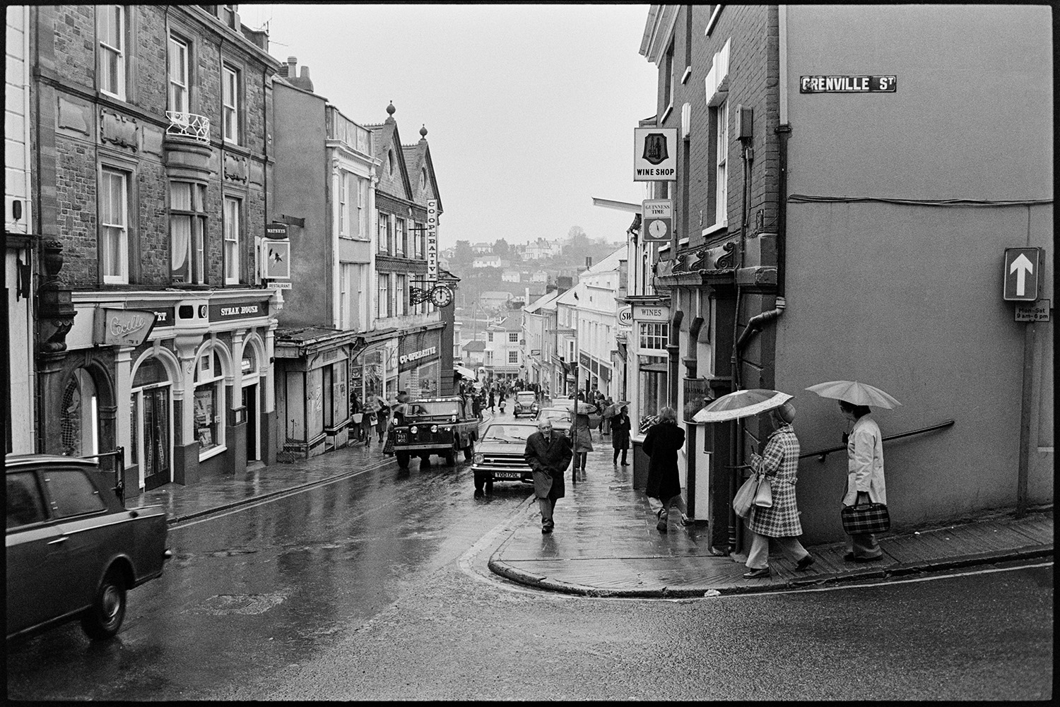 Sign outside Grocery stores in street with passers by. 
[Shoppers and passers by in Buttgarden Street, Bideford. Shop fronts, including the Co-Operative, Wine Shop and Steak House, and cars can also be seen on the street. The road junction and sign for Grenville Street is also visible.]