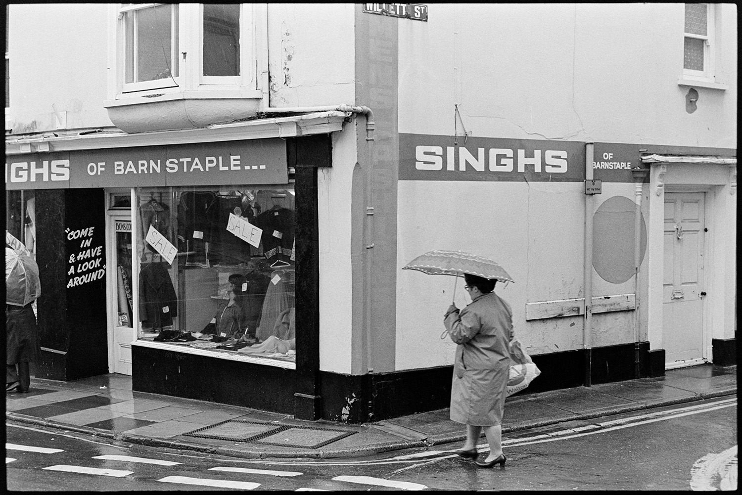 Street scenes, shoppers with umbrellas. 
[A woman holding an umbrella crossing the junction where Willett Street meet Mills Street in Bideford. Singhs of Barnstaple clothes shop is on the corner of the junction.]