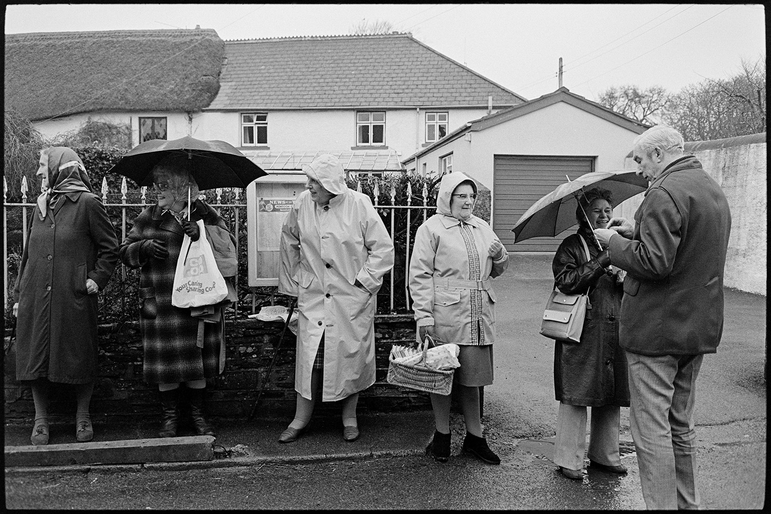 Getting ready for Pancake race in village street and having race. 
[Women and a man lining a street in Dolton to watch the pancake race on Shrove Tuesday. They are wearing rain coats and holding umbrellas.]