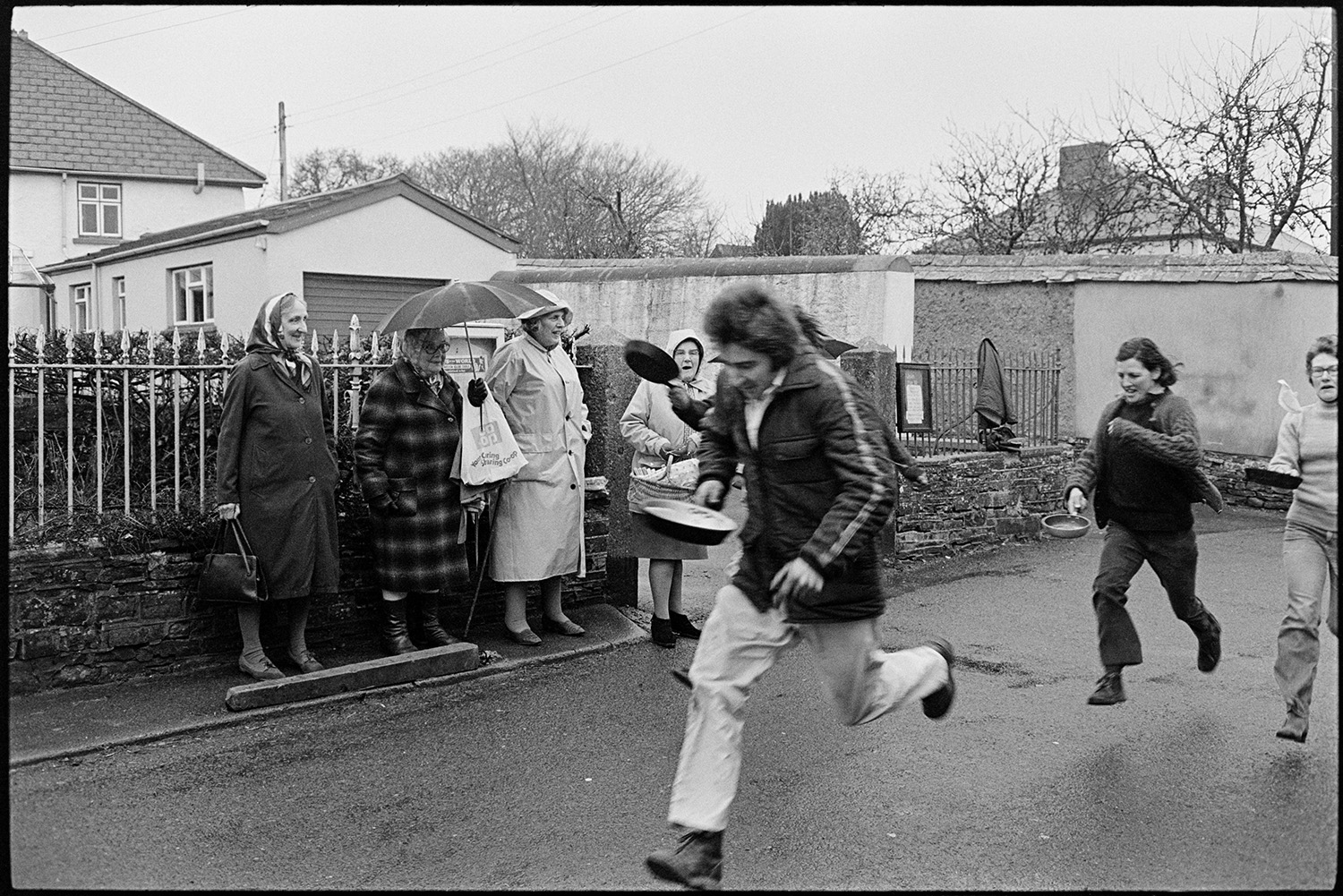 Getting ready for Pancake race in village street and having race. 
[Men and women taking part in a pancake race through a street in Dolton on Shrove Tuesday. A group of women are watching the race from the side of the street.]