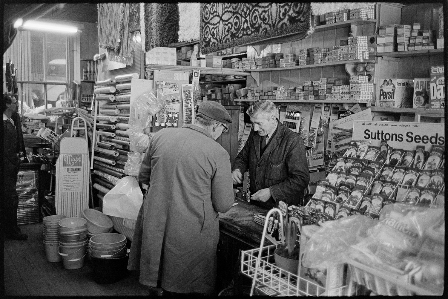 Interior of hardware store with proprietors and customers. 
[The interior of Ellacotts hardware shop in Market Street Hatherleigh. Jim Hockin is serving a customer. Various goods, including Sutton's seeds, buckets and rugs, can be seen in the shop.]