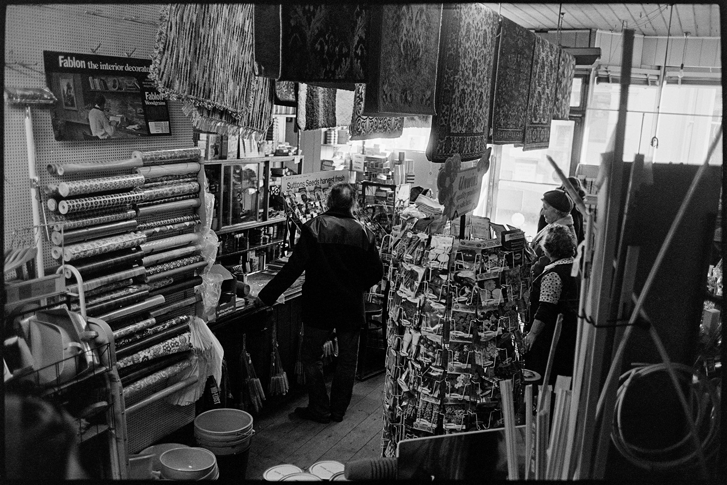 Interior of hardware store with proprietors and customers. 
[Customers queuing at the shop counter in Ellacotts hardware store in Market Street. Hatherleigh. Various goods are on display, including packets of seeds, rugs, buckets and rolls of wallpaper.]