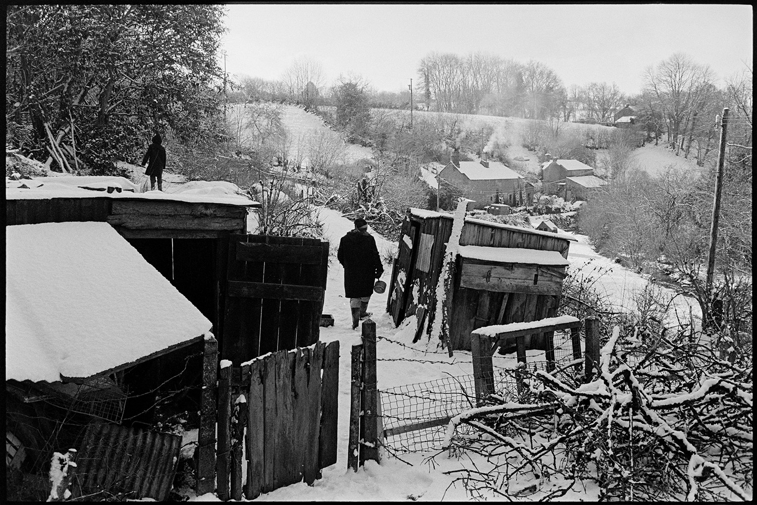 Snow scene with sheds. 
[People checking on livestock, possibly chickens or poultry, in wooden snow covered sheds at Millhams, Dolton. Cottages with smoke rising from their chimneys can be seen in the background.]