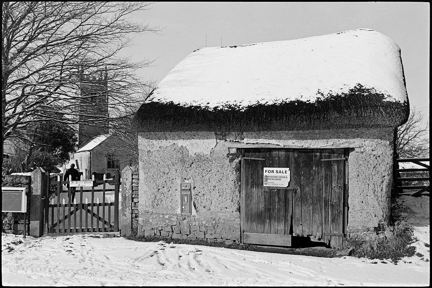 Small cob and thatch stable barn under snow (for sale) now modernised. 
[A cob and thatch barn with a post box in its wall by the church at Dowland. It is covered in snows and has a for sale sign fixed to the wooden door.]