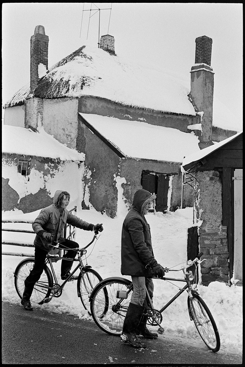Two boys on bicycles in front of snow covered farm. 
[The Lane brothers riding their bicycles past a snow covered thatched farmhouse in Beaford.]
