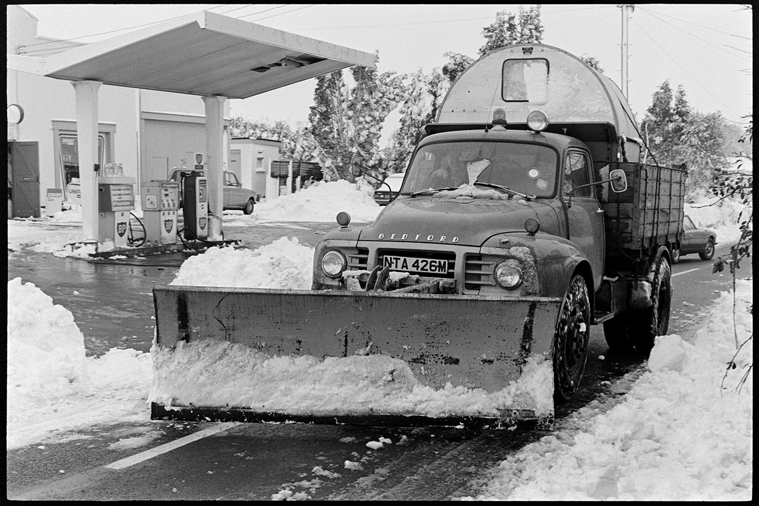 Snow plough going through village. 
[A snow plough passing  garage in Beaford. Mounds of snow can be seen on the side of the road and petrol pumps are visible in the garage.]