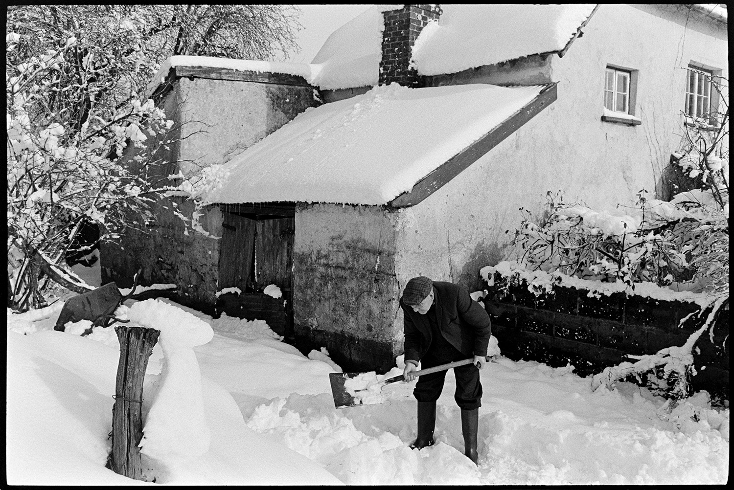 Man clearing snow in front of farmhouse. 
[A man, possibly Cyril Bennett, clearing snow using a shovel, in front of the farmhouse at Cuppers Piece, Beaford.]