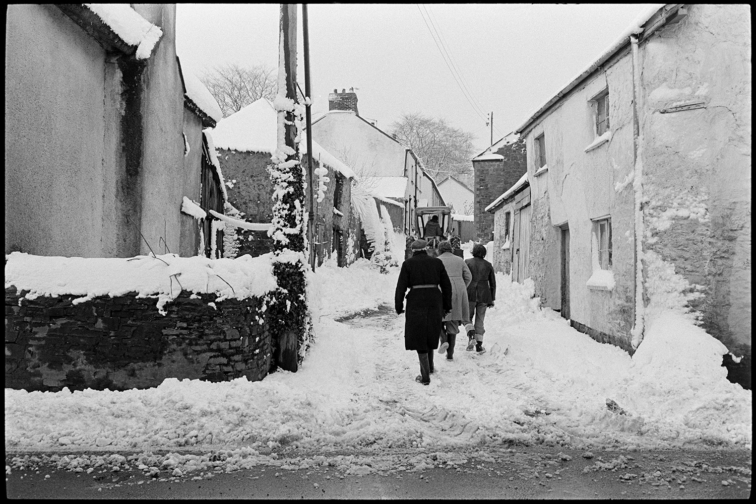 Snow, street scene with people clearing up. 
[People going to clear snow from a street in Beaford. A tractor can be seen further up the street.]