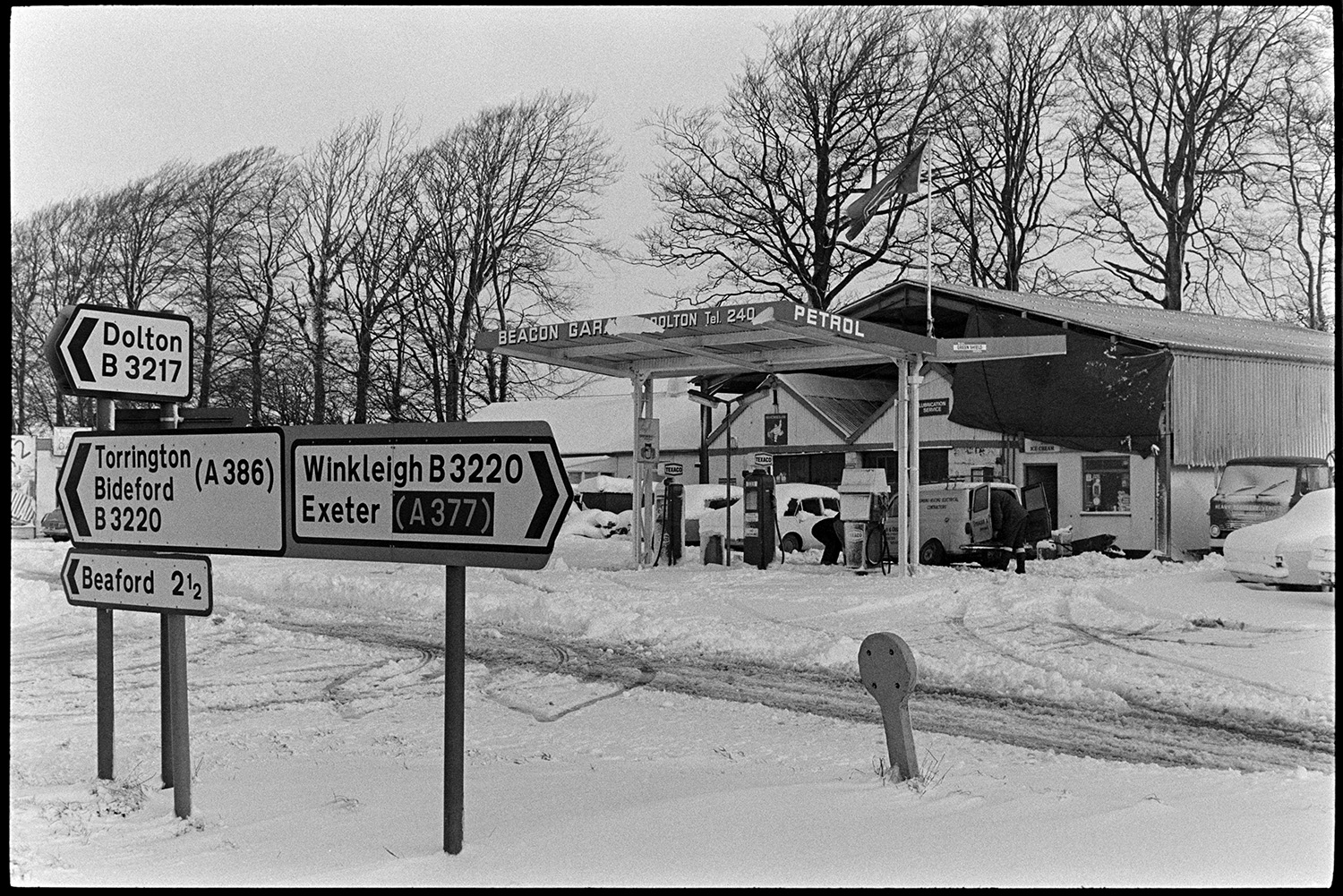 Road signs and garage in snow. 
[Road signs on the snow covered road outside Beacon Garage at Dolton. A van is at the garage behind the petrol pumps.]