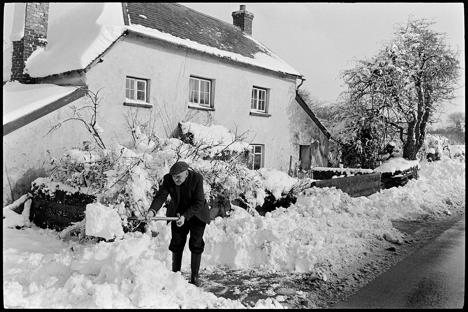 Man clearing snow in front of farmhouse. 
[A  man, possibly Cyril Bennett, clearing snow using a shovel, in front of the farmhouse at Cuppers Piece, Beaford.]
