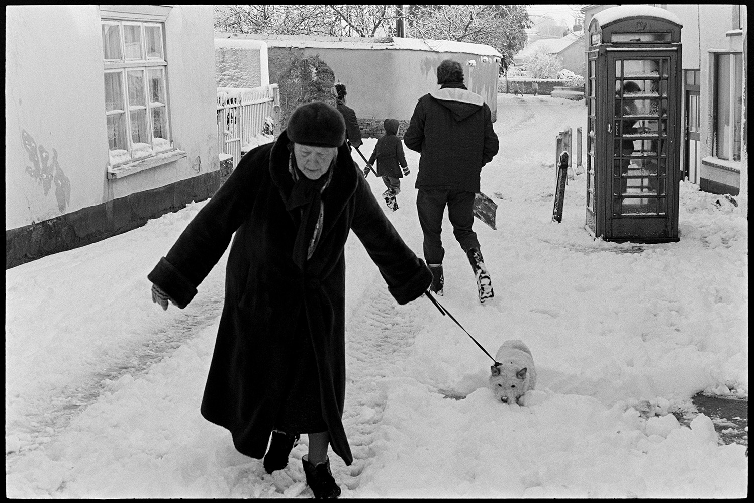 Snow, man taking hay through village to fields, woman with dog. 
[A woman walking her dog through the snow in Fore Street, Dolton. Other people are walking past her in the background, including Cyril Nott carrying a hay bale, and a person is using the telephone box.]