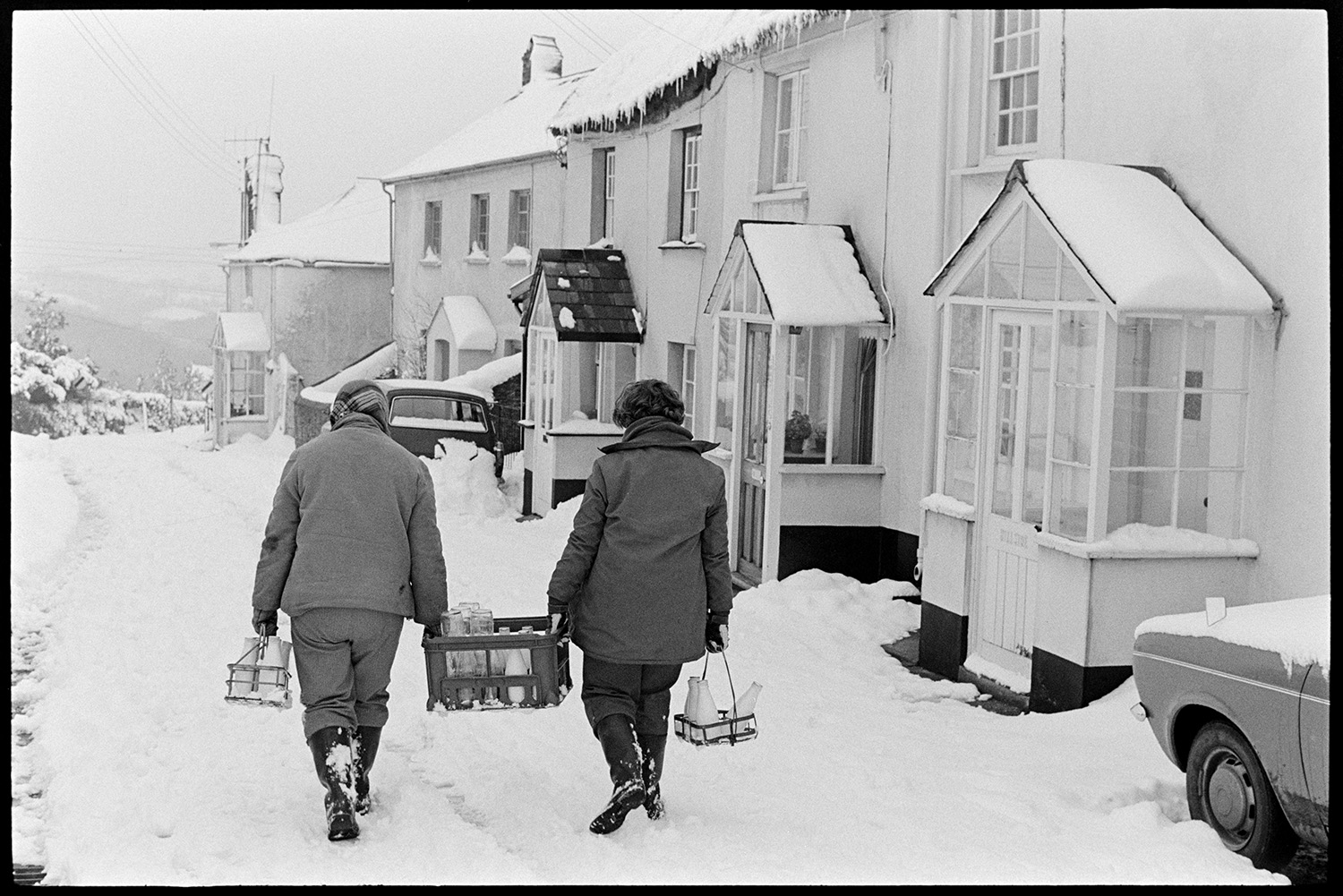Snow, women delivering milk in village, dog. 
[Dorothy Hiscock and her daughter, Diane Hiscock, delivering milk to houses in West Lane, Dolton. They are carrying a crate of milk bottles between them and another holder of milk in their other hand. The lane and houses are covered in snow. Icicles are hanging from some of the roofs.]