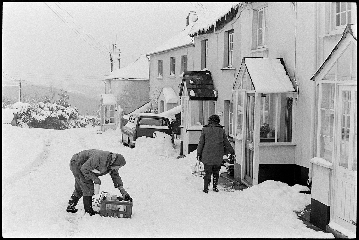 Snow, women delivering milk in village, dog. 
[Dorothy Hiscock and her daughter, Diane Hiscock, delivering milk to a house in West Lane, Dolton. The lane is covered with snow and the porch of one of the houses also has snow on its roof. Icicles are hanging from some of the roofs.]