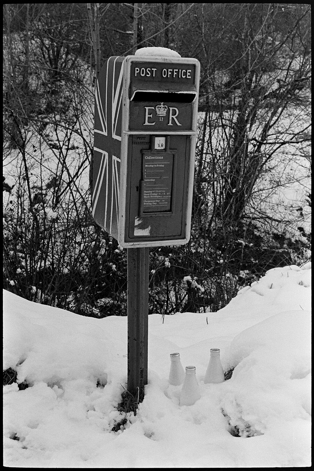 Letter box in snow, painted with flag for Jubilee. 
[A post box covered in snow at Woolridge Cross, Dolton. It has been painted with a Union Jack to celebrate the Silver Jubilee of Queen Elizabeth II the year before. Three milk bottles are stood in the snow under the post box.]
