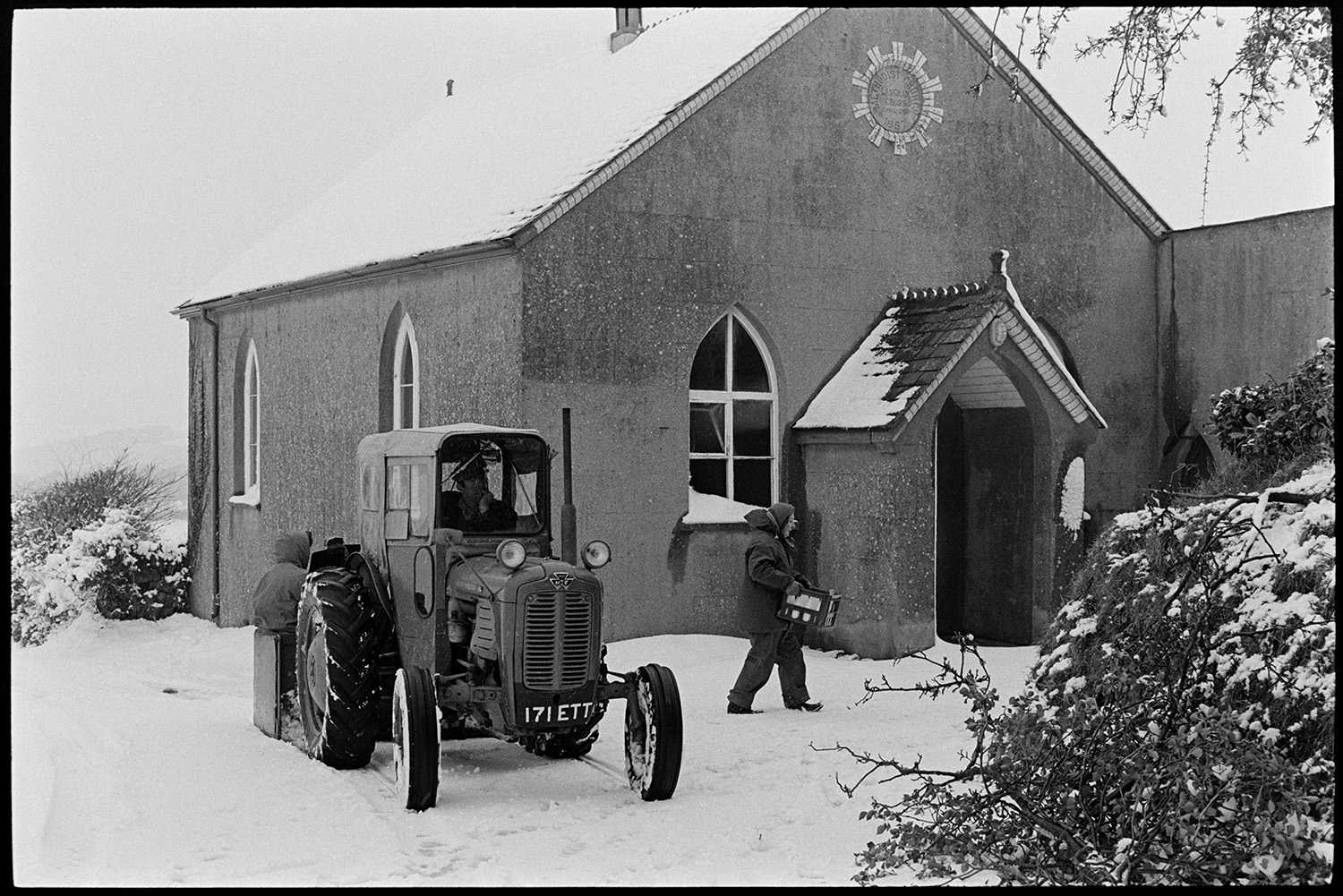 Snow, delivering milk with tractor. 
[Dorothy Hiscock delivering a crate of milk to a building, possibly a chapel, at Langham, Dolton. A tractor is transporting her on the milk round and a person can be seen sat in the link box attached to the tractor.]