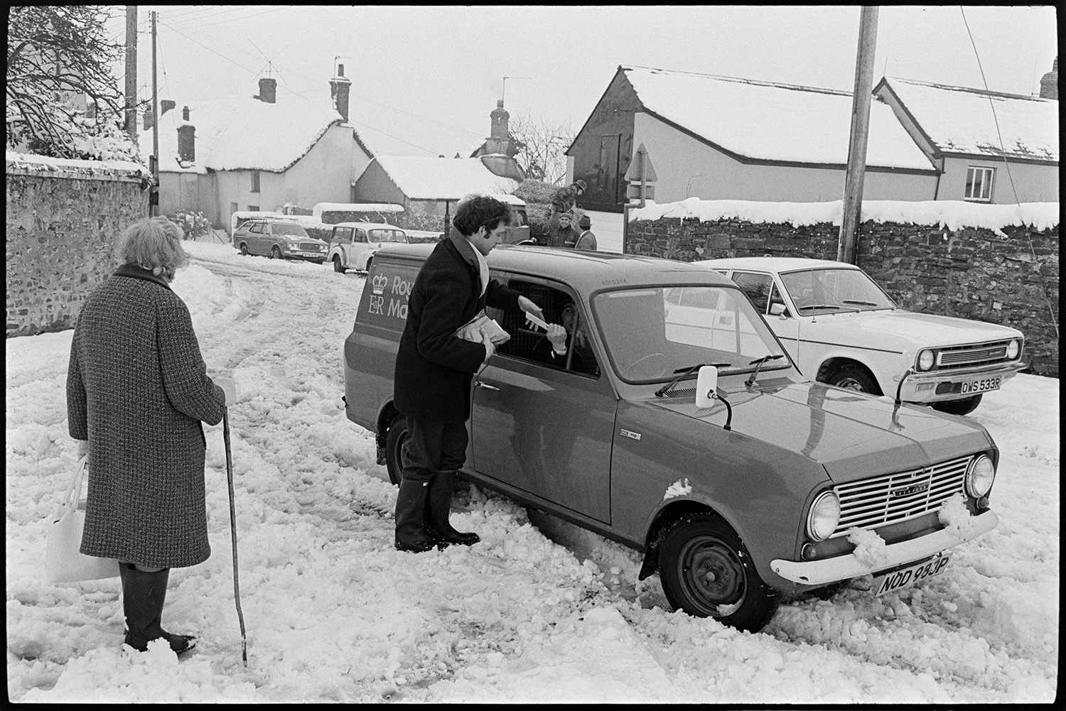 Snow, street scenes, postman handing out mail, shoppers, etc. Tractor and link box. 
[A Royal Mail post van in a snowy street in Dolton. The person in the van is handing a letter to a man, and a woman is watching on. Houses and parked cars covered in snow can be seen in the background.]