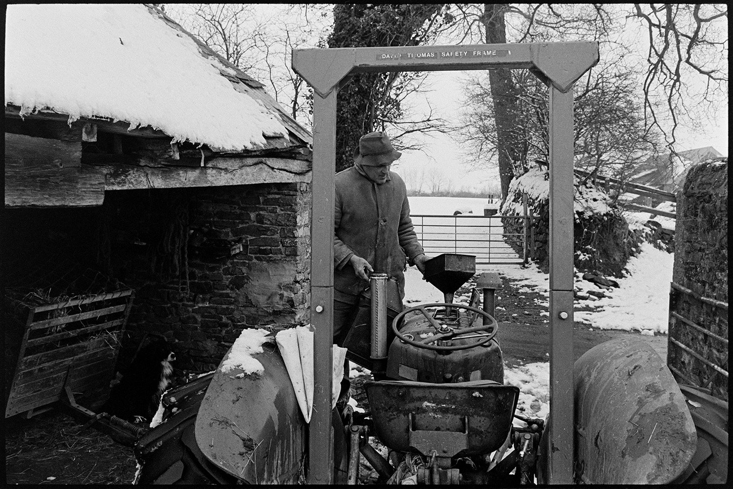 Snow, tractor in farm yard and farmer filling fuel tank with diesel. 
[Alf Pugsley filling his tractor with diesel in the snowy farmyard at Lower Langham, Dolton. A dog is sat in a barn nearby watching him.]