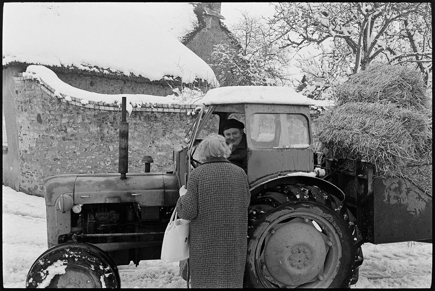 Snow, street scenes, postman handing out mail, shoppers, etc. Tractor and link box. 
[A woman talking to Dennis Harris in a tractor on a snowy road in Dolton. A wall and thatched cottage covered in snow are visible in the background.]