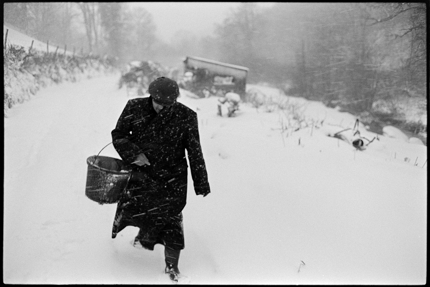 Snow, farmer returning from feeding animals in shed. 
[Ivor Brock walking along a snow covered lane in a blizzard at Millhams, Dolton. He is carrying a bucket after feeding livestock in the shed in the background.]
