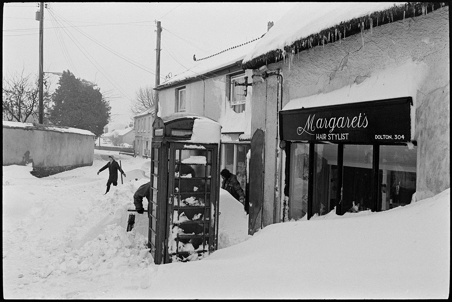 Snow, snowdrifts in village after blizzard, cars, people shovelling, telephone kiosk. 
[People shovelling snow in Fore Street, Dolton by the telephone box and Margaret's hairdressers. Icicles are hanging from the roof.]