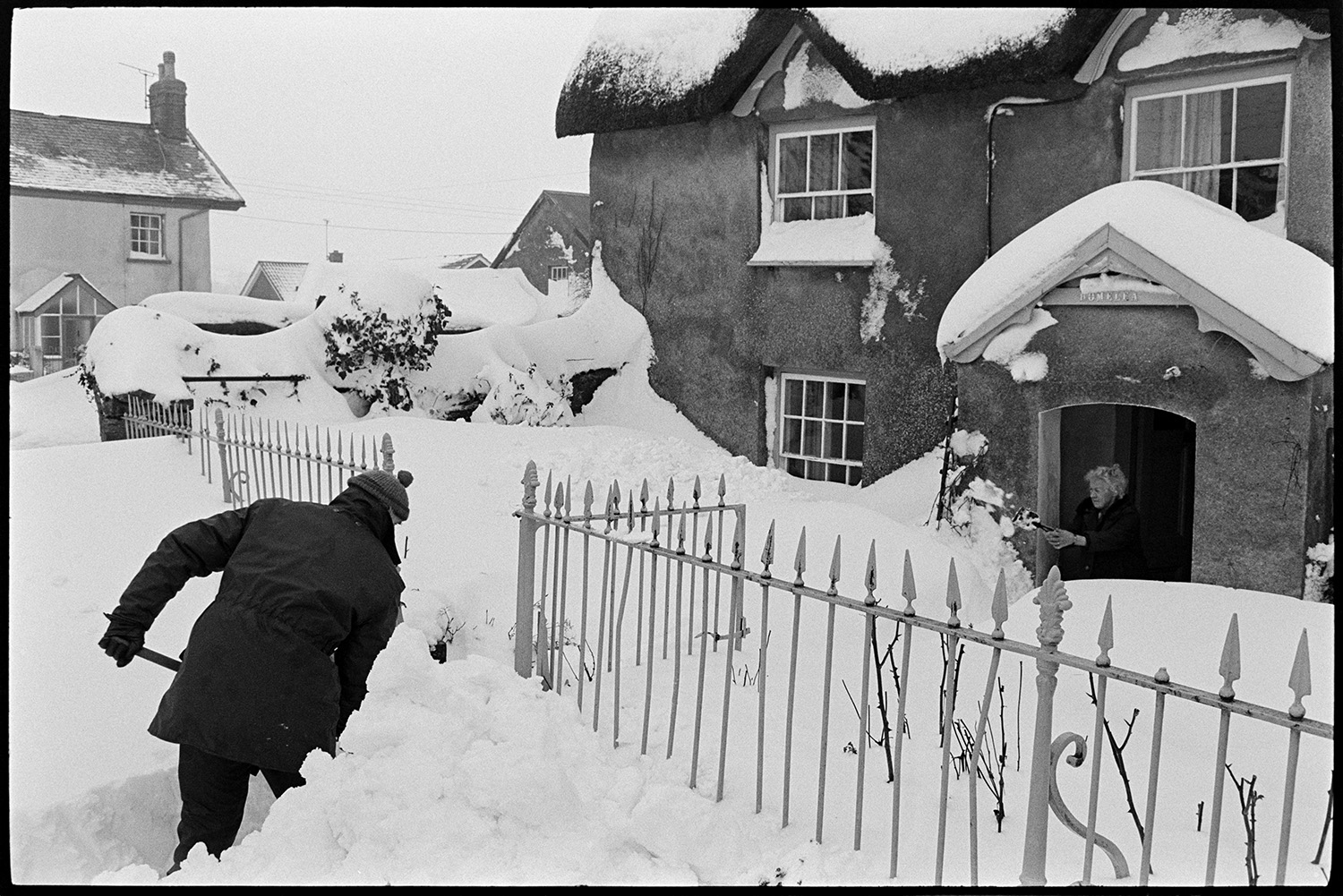 Snow, snowdrifts in village after blizzard, cars, people shovelling, telephone kiosk. 
[Derek Marden shovelling snow outside Homelea cottage in Dolton. A woman is shovelling snow by the porch of the cottage. The snowdrifts are against the railings and ground floor windows of the cottage.]