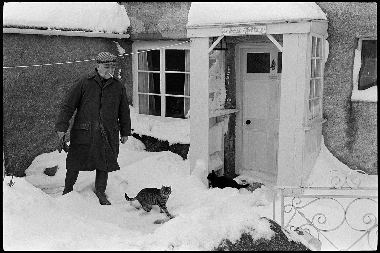 Snow, snowdrifts in village after blizzard, cars, people shovelling, telephone kiosk. 
[Alf Folland and a cat in the snow, outside Verbena Cottage, Dolton.]