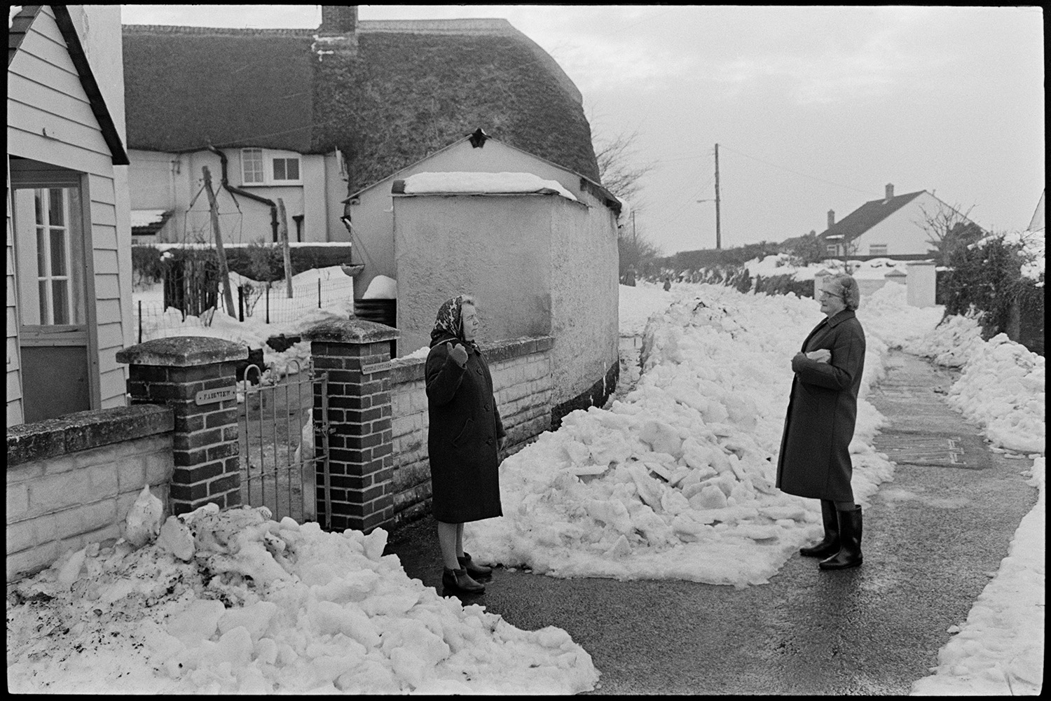 Snow, two women chatting in street. 
[Mrs Friend and another woman talking in the street outside Fair View Cottage in West Lane, Dolton. A pathway has been cleared through the snow in the street.]