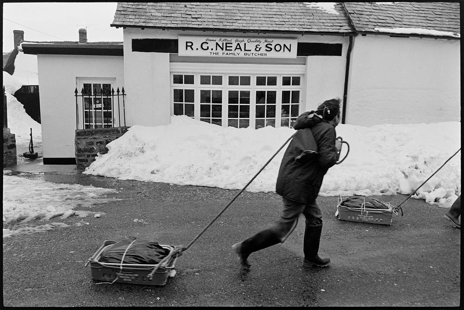 Snow, butcher setting off with meat on sledge for next village, Merton cut off. 
[Bill Davey, butcher, and another person dragging sledges with meat past R G Neal & Son butcher's shop in Dolton. They are going to deliver the meat to Merton which was cut off in the snow.]