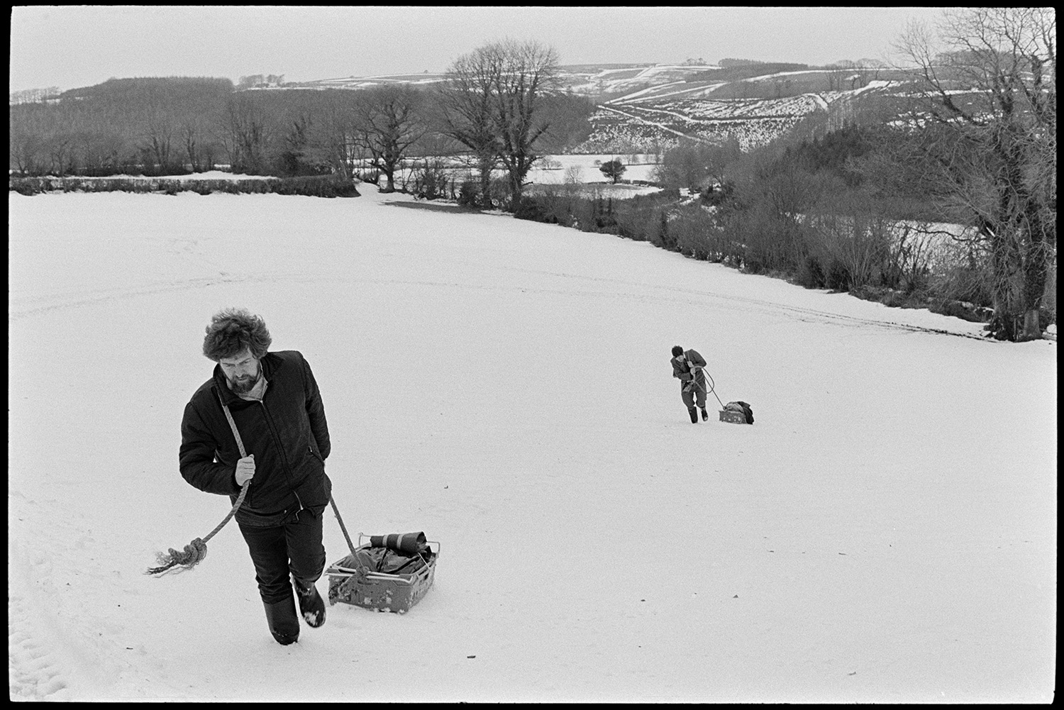 Snow, butcher setting off with meat on sledge for next village, Merton cut off. 
[Bill Davey and another man dragging sledges with meat through a snow covered field at Langham, Dolton. They are going to deliver the meat to Merton which was cut off in the snow. A landscape of snow covered fields and trees can be seen in the background.]