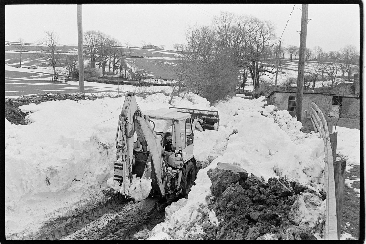 Snow, men clearing roads with digger, JCB. 
[A JCB digger clearing snowdrifts at Dolton. Fields and trees can be seen in the background.]