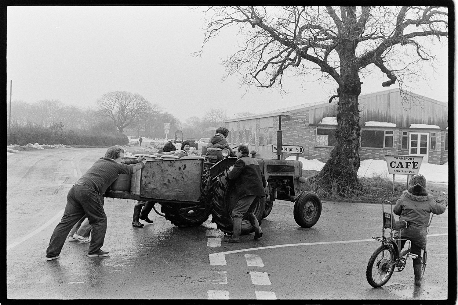 Snow, men pushing tractor. 
[Men pushing a tractor and link box, filled with milk churns, along a road in Winkleigh. Two boys re riding past on bicycles. A sign on the side of the road is pointing to the Transport Café. Snow is piled up on the side of the road.]