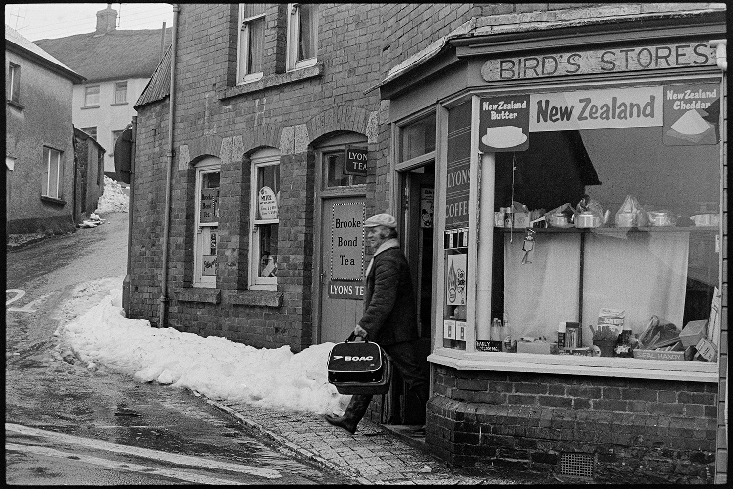 Snow street scenes with shops and shoppers.
[Man with shopping bags stepping into a snow lined street outside Bird's Stores in Winkleigh. Teapots are displayed in the shop window.]