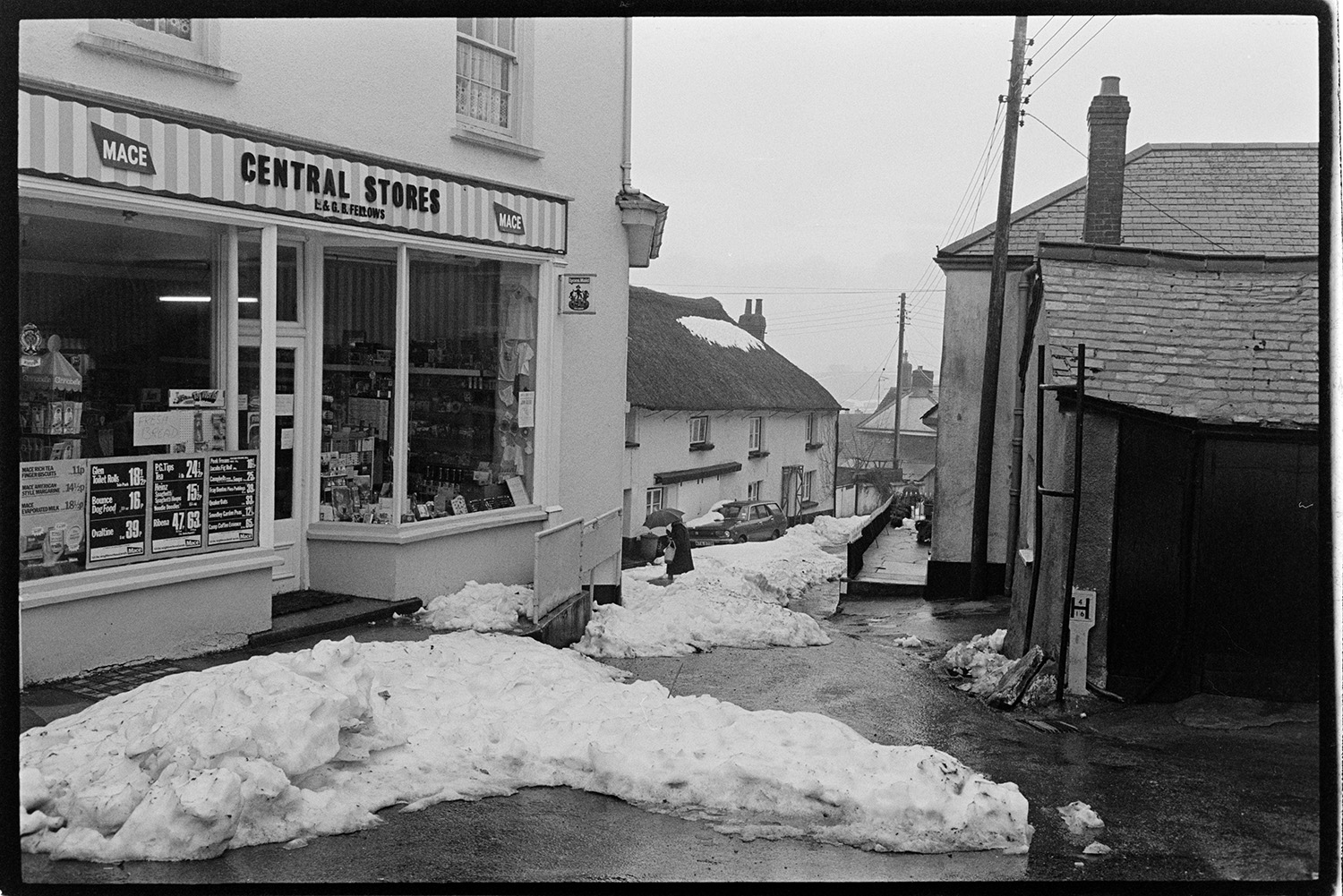 Snow. Street scenes with shops and shoppers.
[Snow in the street outside Central Stores, Winkleigh. A person walking in the snow with an umbrella past a thatched cottage can be seen further down the street.]
