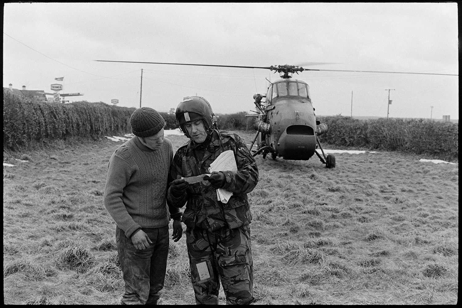 Snow. Helicopter landing to get directions!
[Helicopter pilot talking to a man in a field at Dolton Beacon to get directions. The helicopter is in the background and a Texaco garage sign can be seen over the hedge pf the field.]