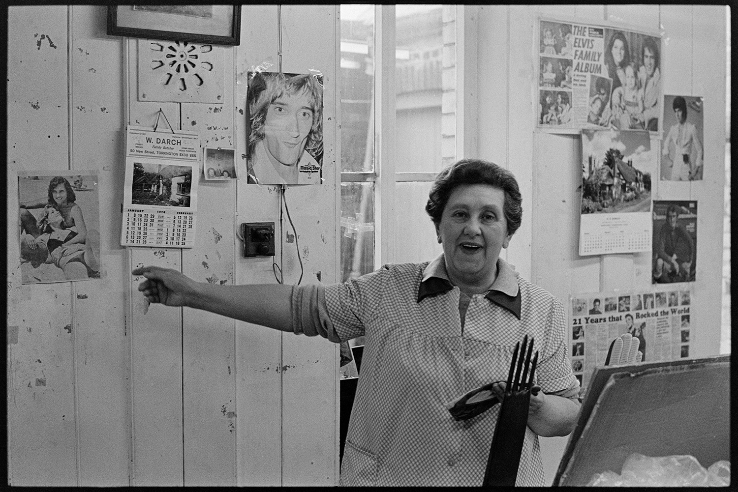 Workers in glove factory. Machinery. Pin-ups!
[A woman pointing to pictures, posters and calendars on the wall at Vaughans Glove Factory, Halsdon Terrace, Torrington. The posters include pictures of Elvis and Rod Stuart.]