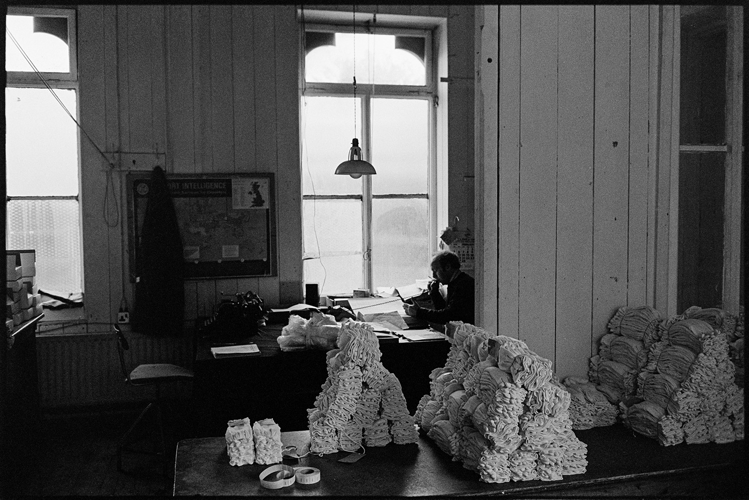 Workers in glove factory. Old machinery. Canteen. Office with man on telephone. Deliveries.
[Piles of gloves on a table with a man working in the office in the background at Vaughans Glove Factory, Halsdon Terrace, Torrington.]