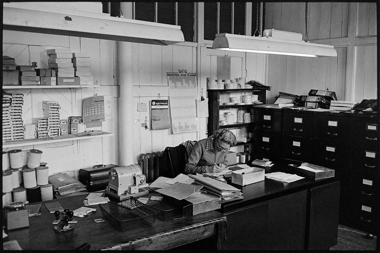 Workers in glove factory, old machinery, canteen, office with man on telephone, deliveries. 
[A woman working at a desk with paperwork at Vaughans Glove Factory , Halsdon Terrace, Torrington. Filing cabinets can be seen in the background.]