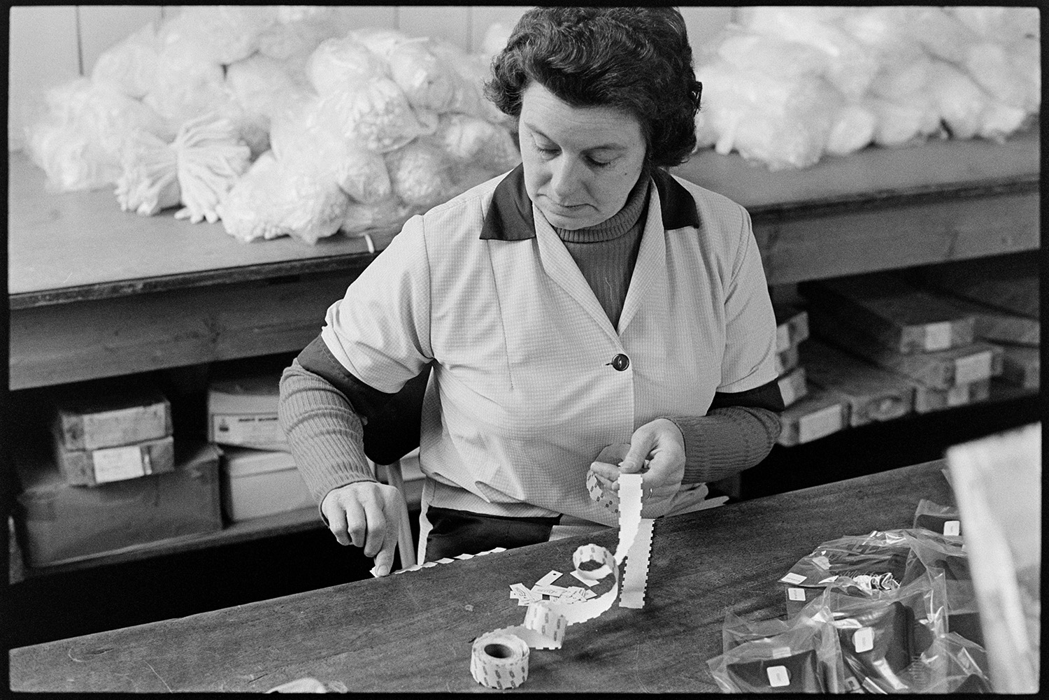 Workers in glove factory, old machinery, canteen, office with man on telephone, deliveries. 
[A woman working at Vaughans Glove Factory, Halsdon Terrace, Torrington. She is labelling packets of gloves.]