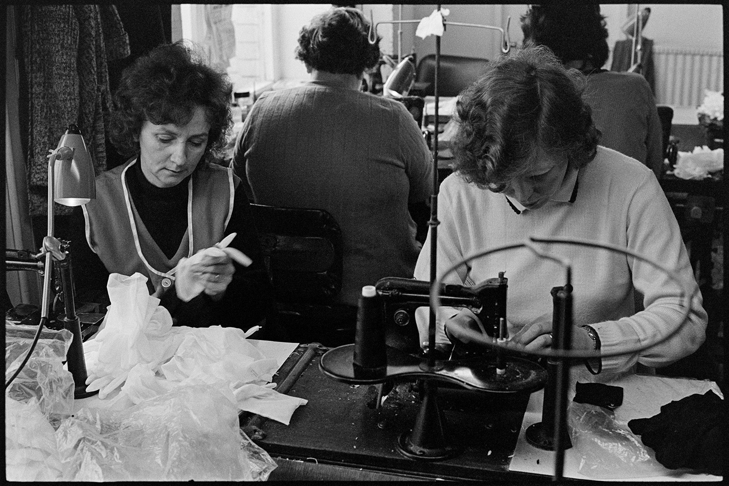 Workers in glove factory, machines and elderly woman at roll top desk doing accounts. 
[Women sat working at sewing machines making gloves at Vaughans Glove Factory, Halsdon Terrace, Torrington.]