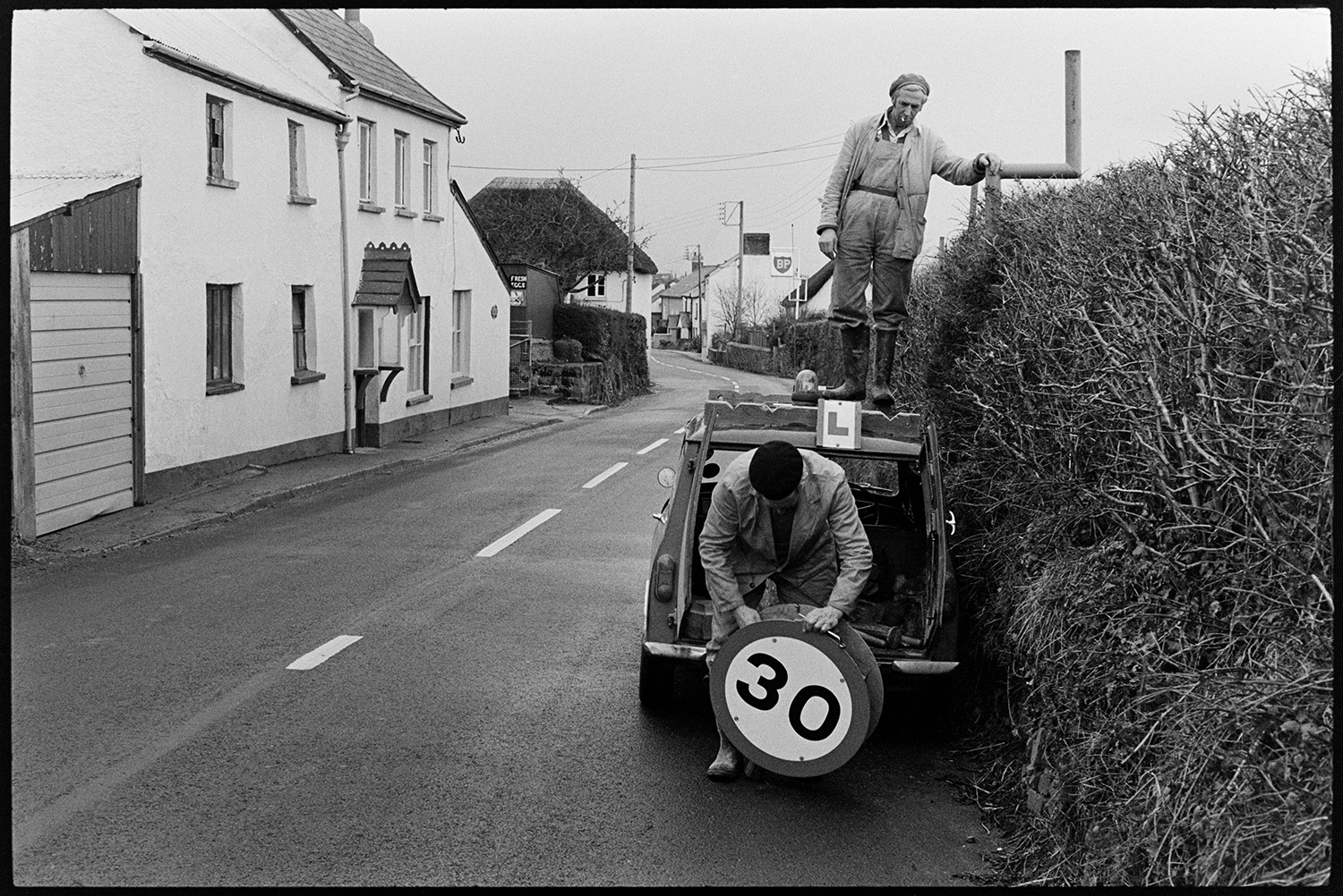 Men putting up 30 mph sign in hedge outside village. 
[Two men putting up a 30 mph road sign in a hedge in Beaford. One of the men is stood on top of a van to put up the sign. Houses can be seen in the background further along the street.]