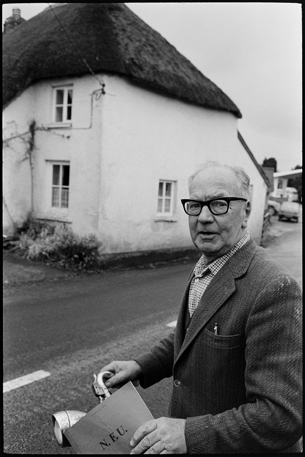 Man standing in road. 
[Philip Heard standing with a bicycle in a street opposite a thatched cottage in Beaford.]