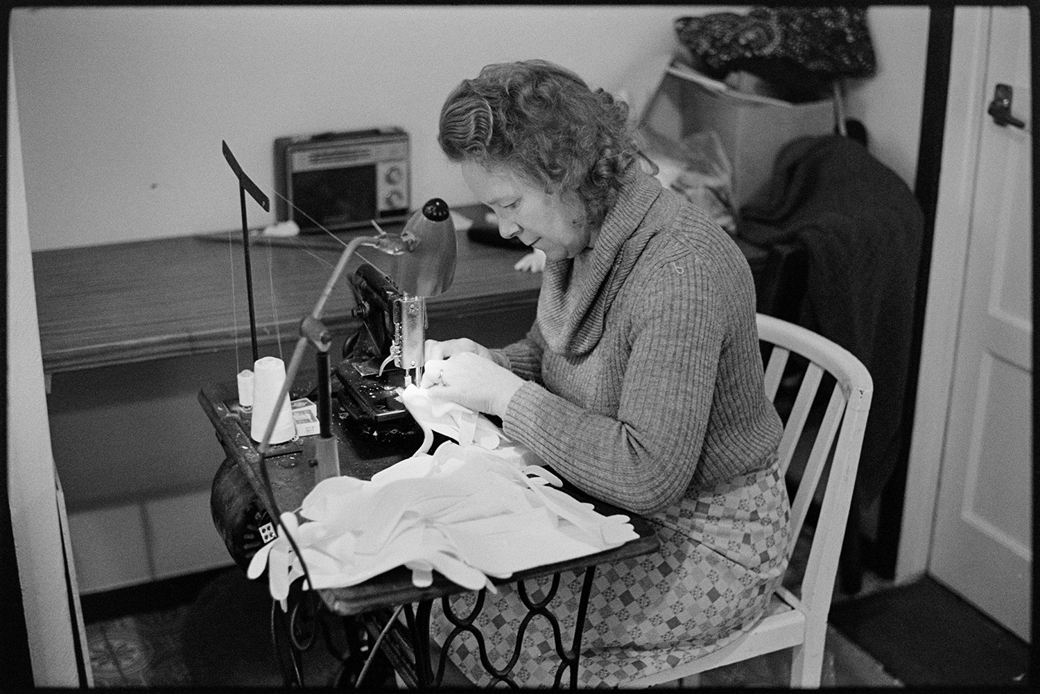 Woman out-worker making gloves at home. 
[Mrs Thacker making sat at a sewing machine making gloves for Vaughans Glove Factory in her home in Torrington.]