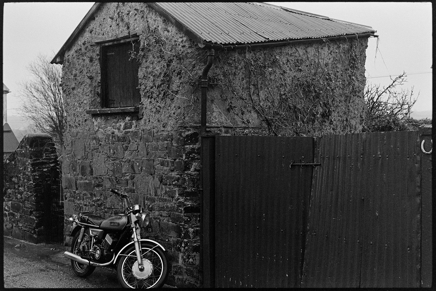 Motorbike parked beside barn shed made of cob with tallet. 
[A motorcycle parked outside a stone and cob barn with a corrugated iron roof in Aller Road, Dolton. A wooden door to the barn tallet is also visible.]