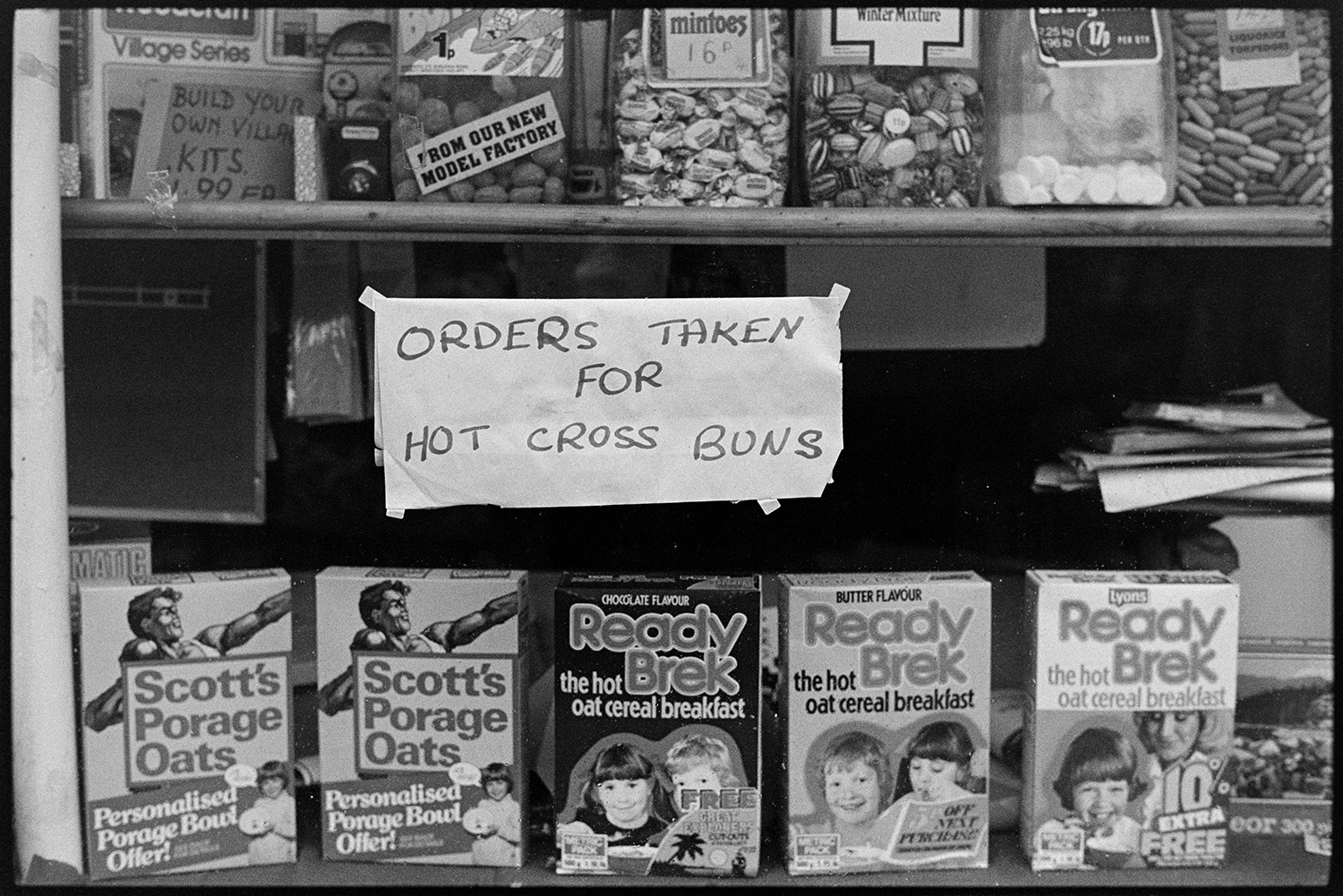 Grocery shop window with price signs and notice, hot cross buns. 
[The shop front window of London House Stores in Fore Street, Dolton. A display of sweet jars, porridge boxes and a sign reading 'Orders Taken for Hot Cross Buns' are visible.]